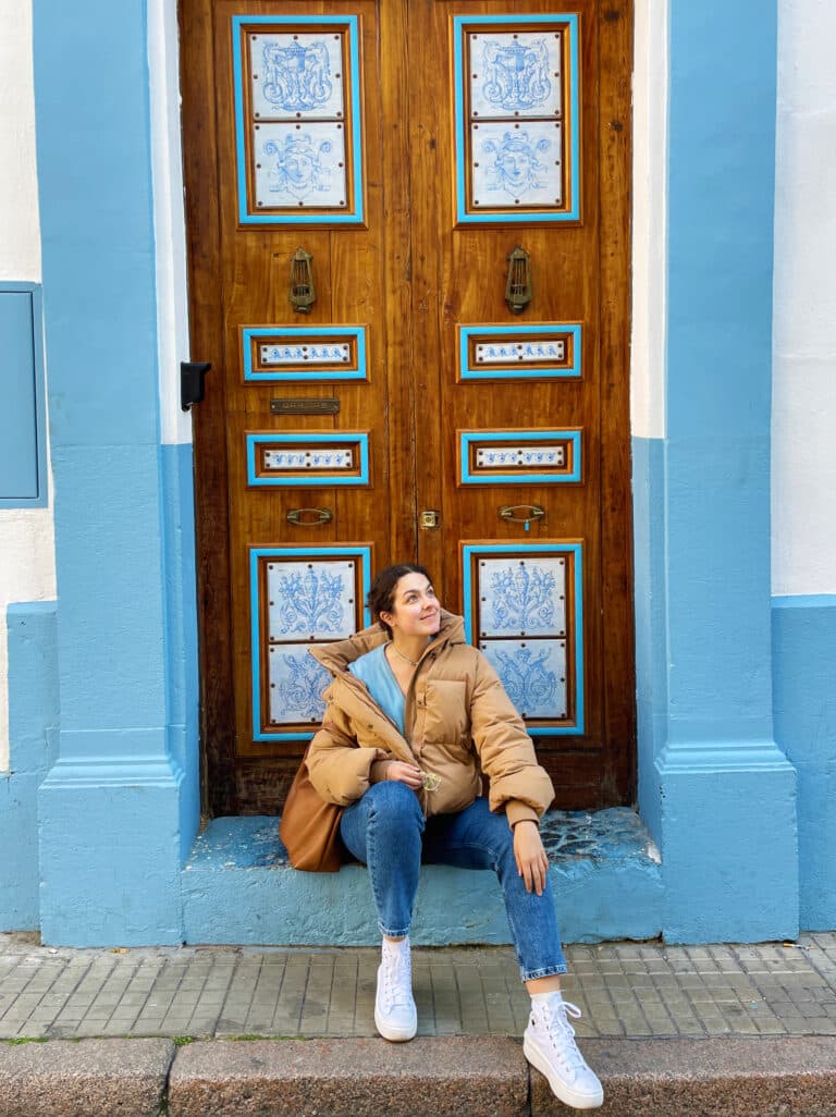 A woman in a tan puffer jacket, blue jeans, and white sneakers sits on steps in front of an ornate wooden door with blue and white tile accents, framed by a light blue wall. She looks to the side and smiles.
