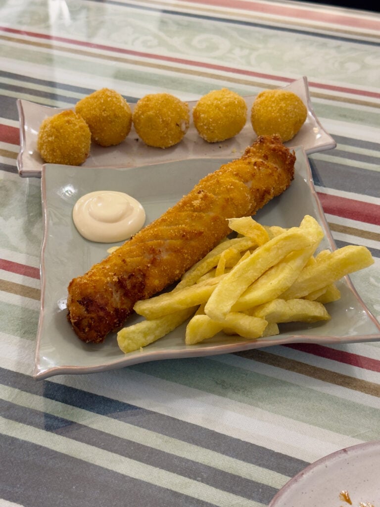 A plate with crispy fries, a breaded fish fillet, five round croquettes, and a dollop of mayonnaise sits on a striped tablecloth.