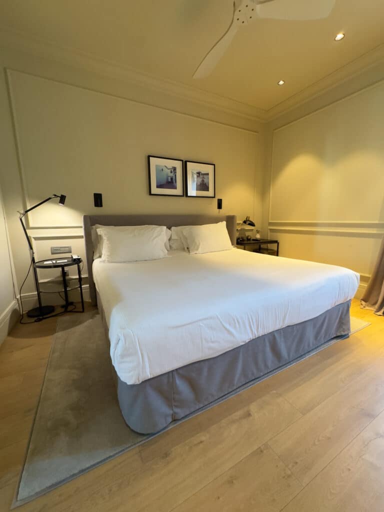 A modern bedroom with a large bed covered in white linens, flanked by two bedside tables with lamps. Two framed pictures hang above the bed. The room has wooden floors, light-colored walls, and a ceiling fan.