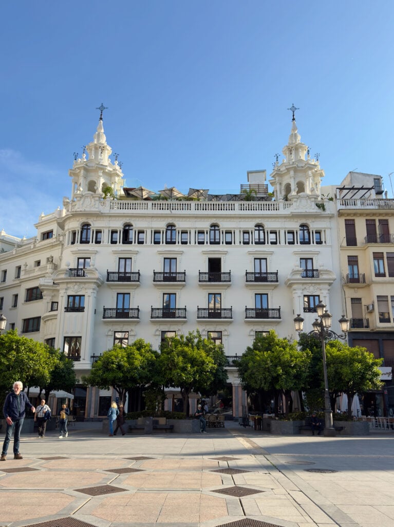 A grand white building with decorative towers and wrought-iron balconies stands behind a row of green trees in a sunny plaza, with a few people walking and relaxing in the square.