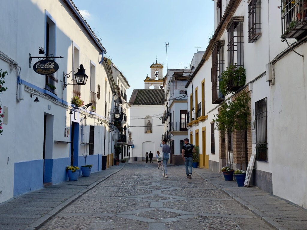 Sunlit cobblestone street in a whitewashed town with blue accents, wrought-iron balconies, and people strolling in the distance.