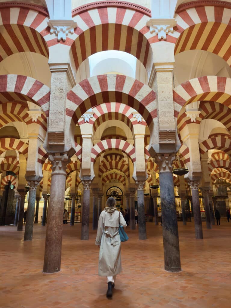 A person in a beige trench coat walks through a hall of Moorish arches with red and white stripes and tall columns, inside the Mosque–Cathedral of Córdoba, Spain.
