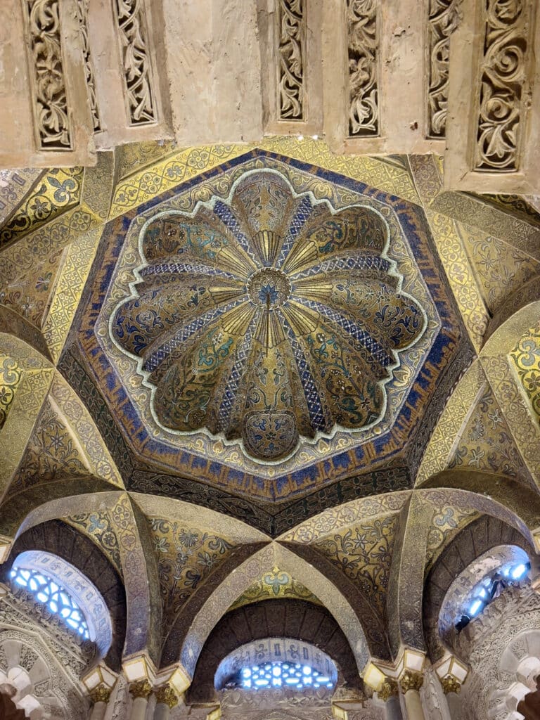 Ornate, colorful dome ceiling with intricate geometric and floral patterns in gold, blue, and brown, surrounded by arches and detailed carvings in a historical building. Light filters through small windows.