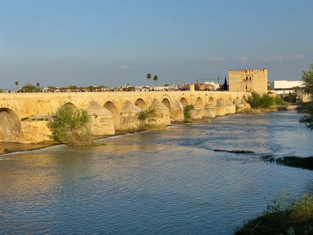 A historic stone bridge with multiple arches spans a calm river under a clear blue sky. On the right, a large square tower stands near the bridge, and small trees grow along the water’s edge.