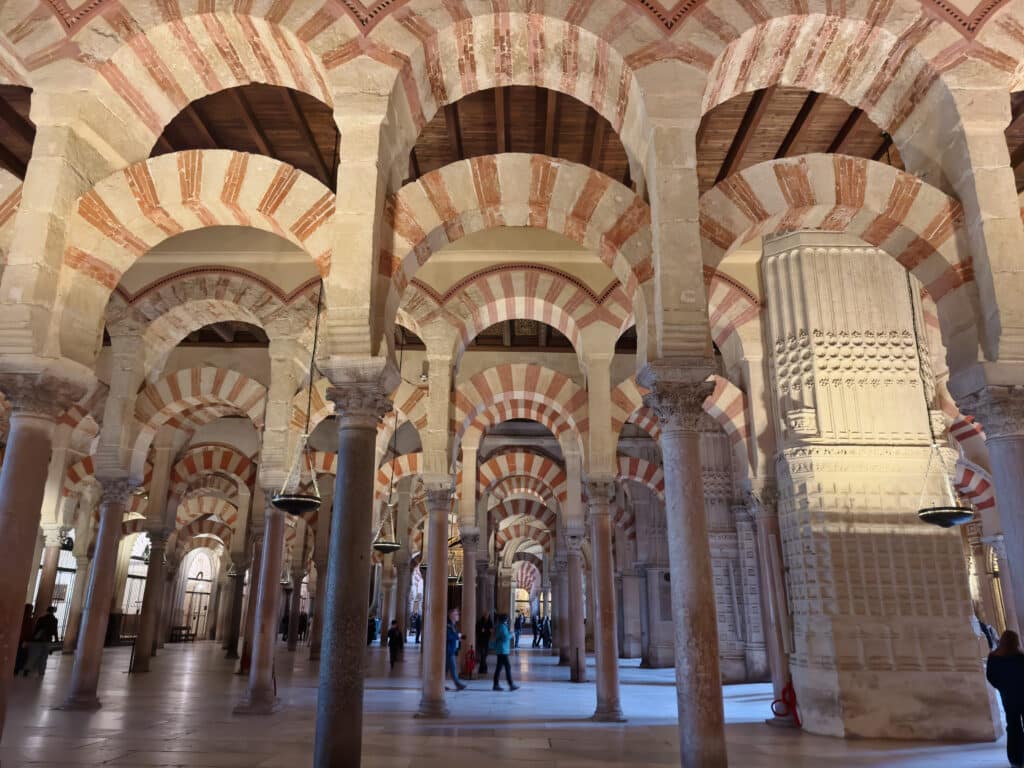 Interior view of the Great Mosque of Córdoba, featuring rows of columns with distinctive red and white striped arches, stone pillars, and a few visitors walking and observing the architecture.