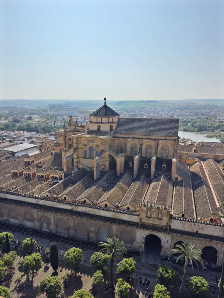 Aerial view of a large historic complex with beige stone walls, a blue-tiled dome, and long sloped tiled roofs, overlooking a town and river in the distance.