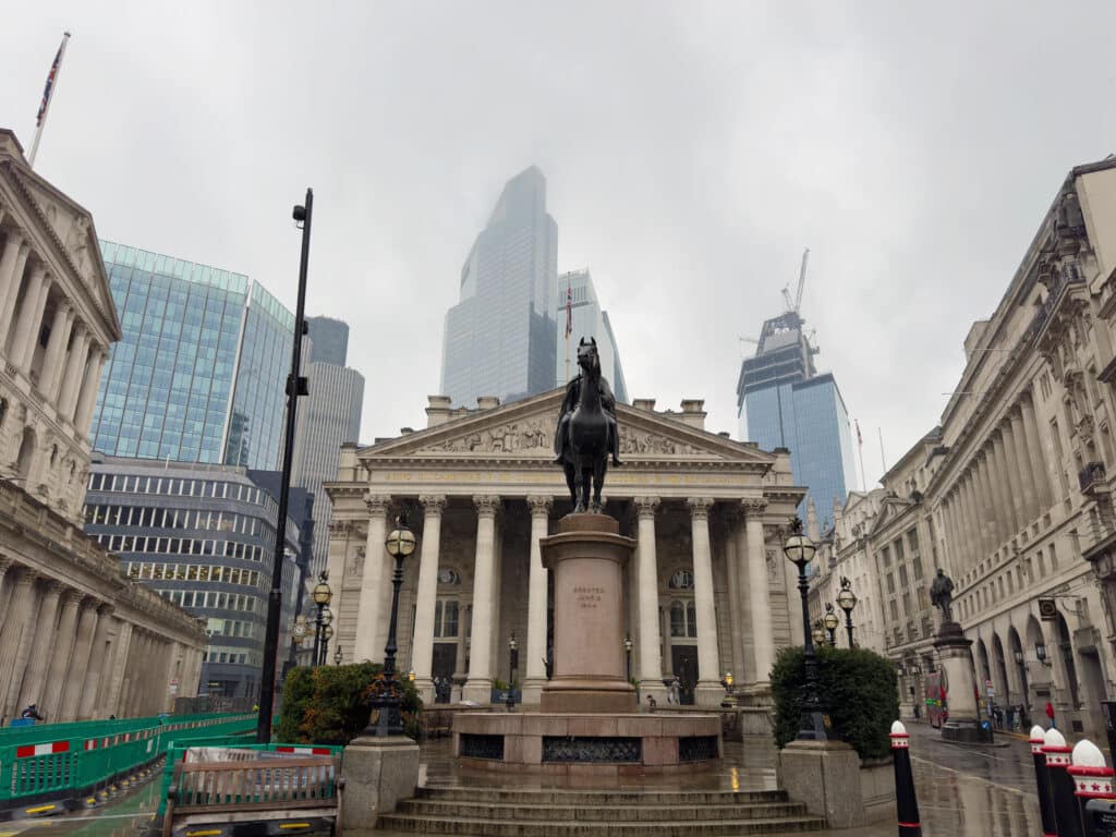 Neoclassical city hall with a statue on a horse in front, modern glass skyscrapers behind, on a cloudy day.