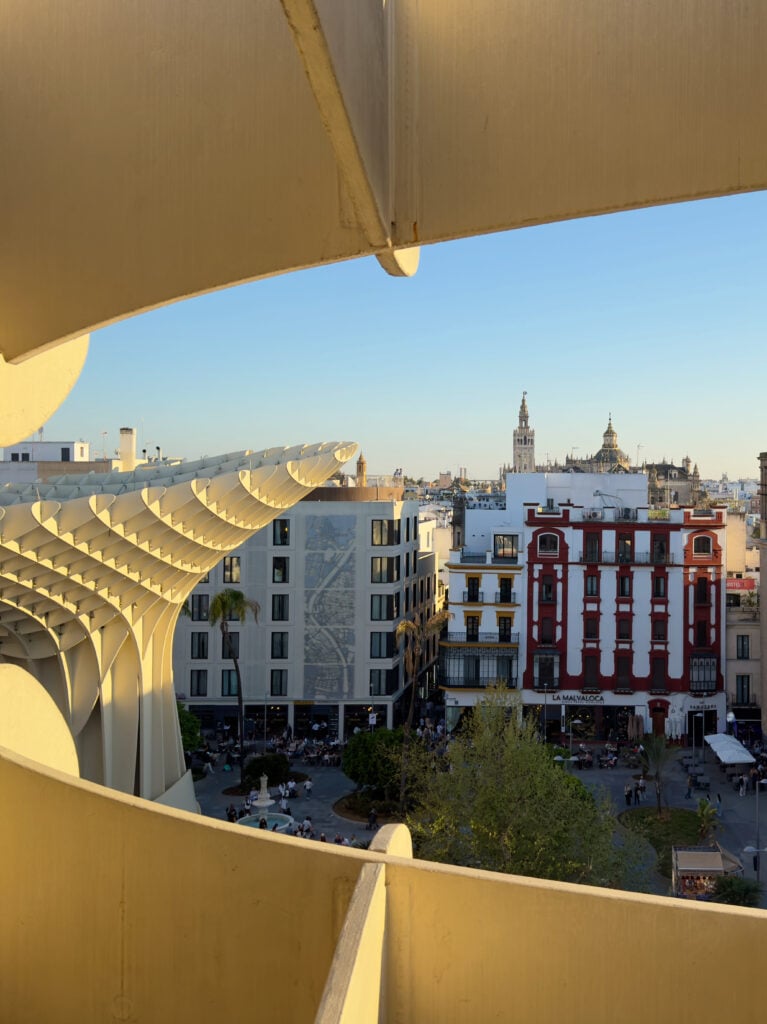 View of a cityscape framed by curving yellow architectural panels, with modern and traditional buildings, a busy plaza, and towers visible in the distance under a clear blue sky.
