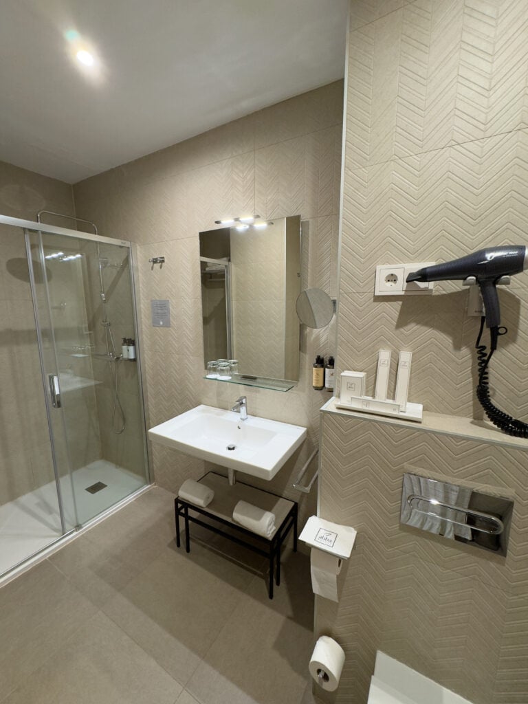 Modern bathroom with beige herringbone walls, a glass shower, wall-mounted sink and mirror, black bench with towels, hairdryer, toiletries, and toilet paper holder. Well-lit and minimalistic design.