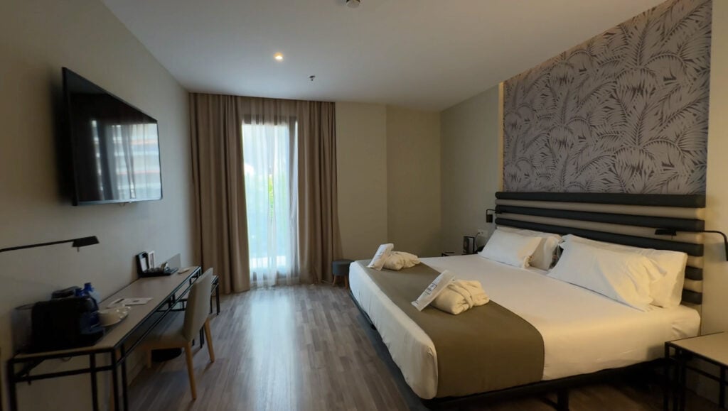 Modern hotel room at abba Sevilla hotel, featuring a large bed, two bedside tables, a wall-mounted TV, a desk with a chair, and a window with sheer curtains letting in natural light—an inviting space that makes this one of the best hotels to stay in Seville near the iconic Setas de Sevilla.