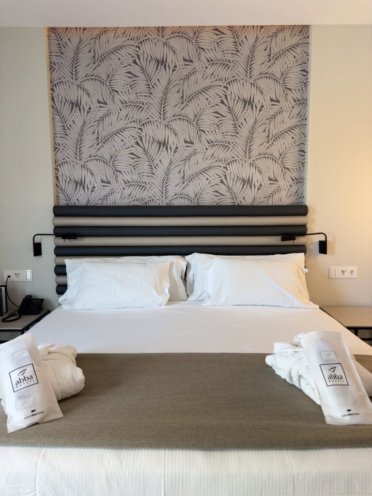 A neatly made hotel bed with two white pillows, a gray headboard, bathrobes, and slippers arranged at the foot. The wall behind features a gray leafy patterned design, reflecting the elegant style of abba Sevilla Hotel—a 4-star hotel often regarded as the best hotel to stay in Seville near Setas de Sevilla and the Real Alcazar.