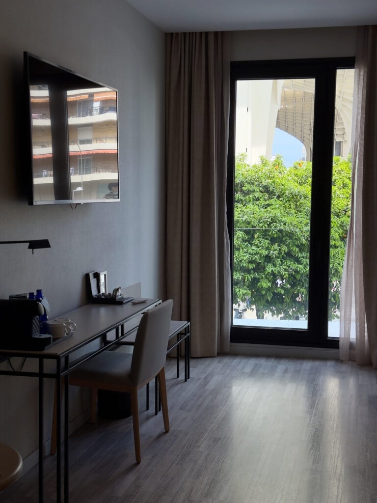 A hotel room with a wall-mounted TV, desk, chair, and large window with curtains. Outside, greenery and part of a building are visible. Located near the iconic Setas de Sevilla, abba Sevilla Hotel is considered one of the best hotels to stay in Seville for both comfort and convenience.