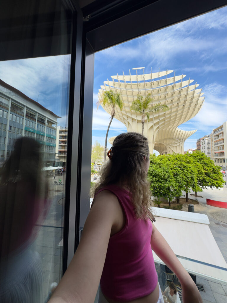 A person, Laura Dolci Travels, in a pink top stands on a balcony at the abba Sevilla Hotel, looking toward the large, modern wooden structure known as Setas de Sevilla (Metropol Parasol), with palm trees and buildings around, under a blue sky with clouds—truly the best hotel to stay in Seville for breathtaking views.