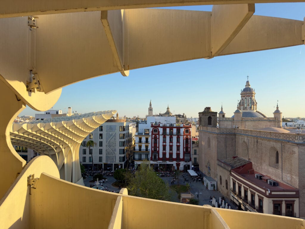 View of Seville cityscape framed by the wooden structure of Metropol Parasol, with historic buildings, a church dome, and the Giralda tower visible under a clear blue sky.