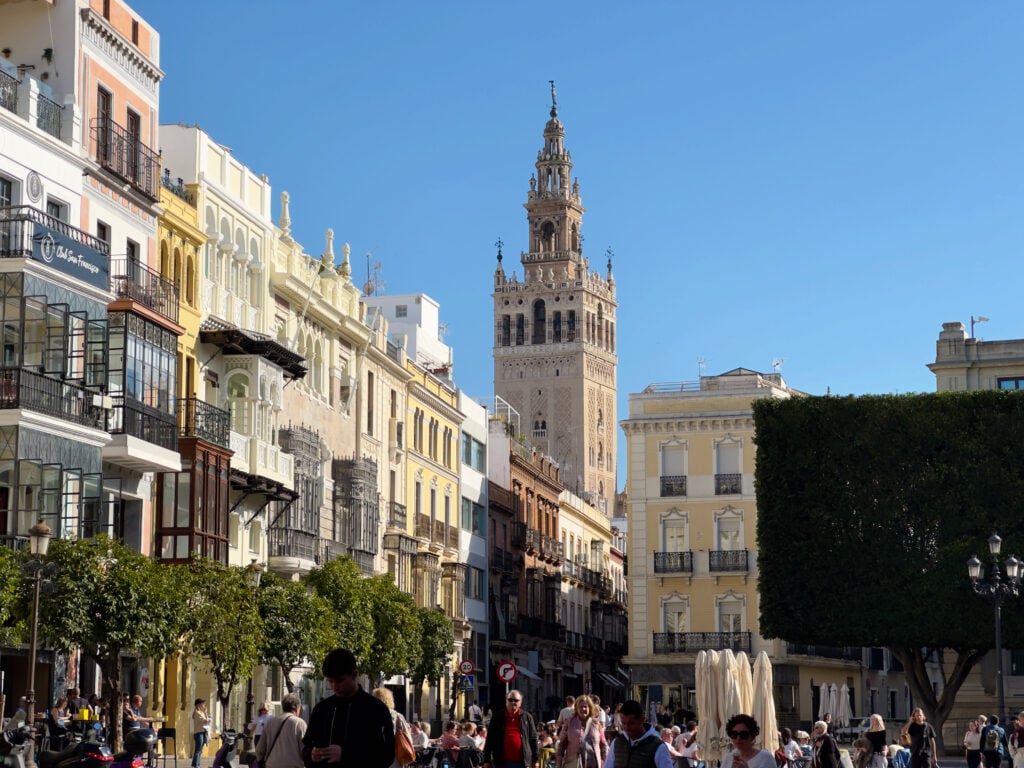 People walk on a sunny street lined with colorful buildings and a tall, ornate tower in the background under a clear blue sky. Trees and a fountain are also visible in the scene.
