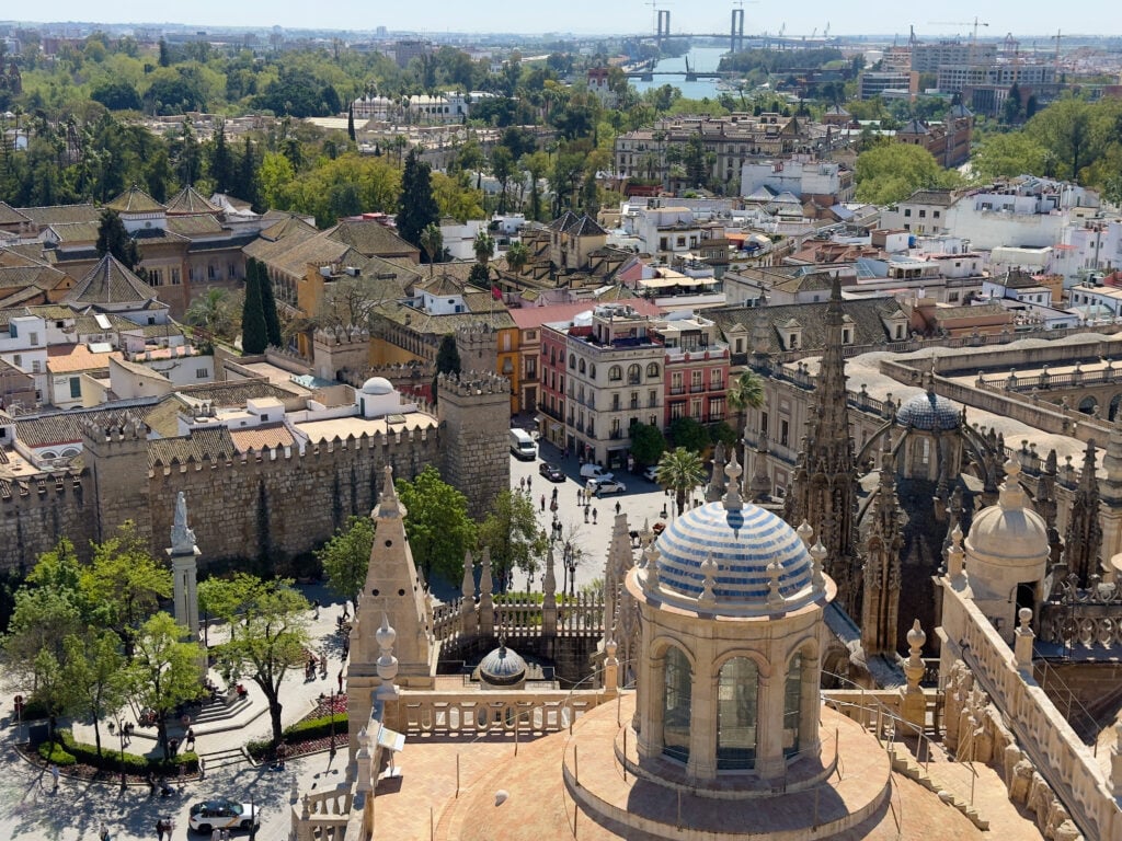 Aerial view of Seville, Spain, showing the Seville Cathedral’s dome, Gothic spires, city buildings in the heart of Santa Cruz, historic walls, trees, and a distant river with a modern bridge under a clear sky.