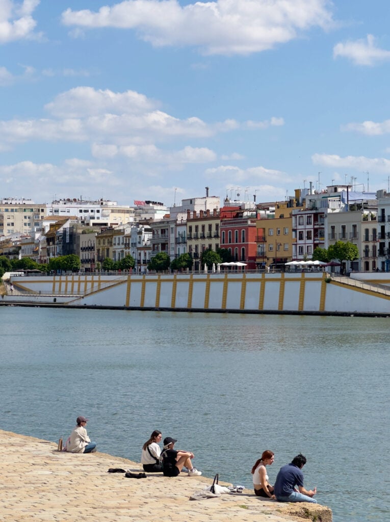 People sit along the Guadalquivir River in Seville, with colorful buildings and the iconic El Arenal neighborhood under a blue sky with scattered clouds.