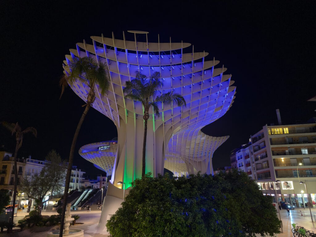 Large, modern wooden structure known as Metropol Parasol, also referred to as the Setas de Sevilla at night, illuminated in blue and green during the Aurora light show, with palm trees and surrounding city buildings visible under a dark sky.