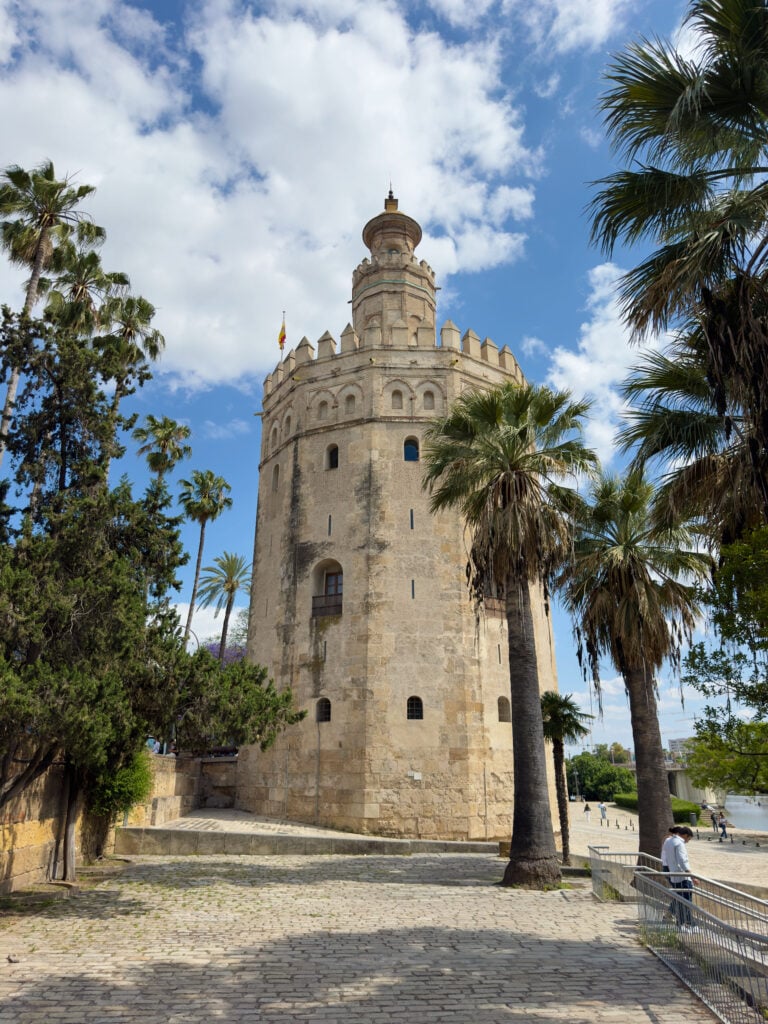 A tall, historic stone tower—the iconic Torre del Oro—stands among palm trees under a partly cloudy sky in Seville. A cobblestone path winds nearby, with people walking in its shade.