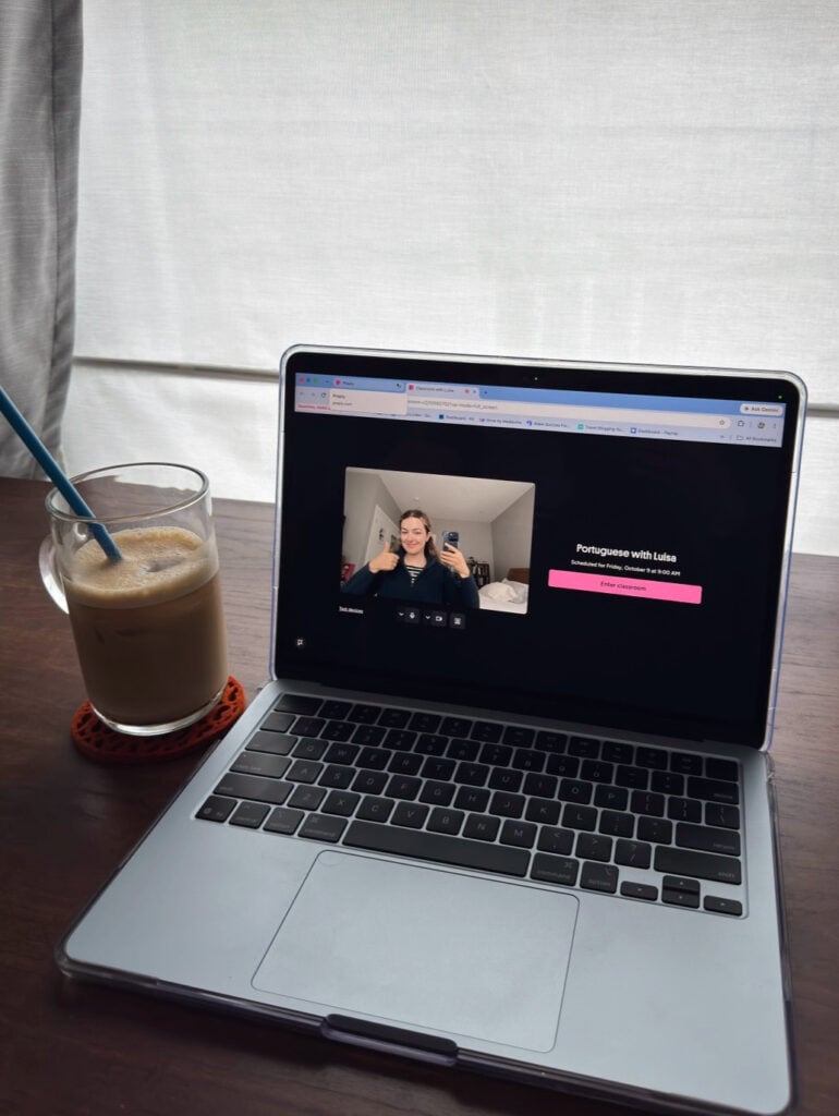 A laptop screen displaying a Preply classroom session with a teacher, next to a glass of iced coffee on a desk.