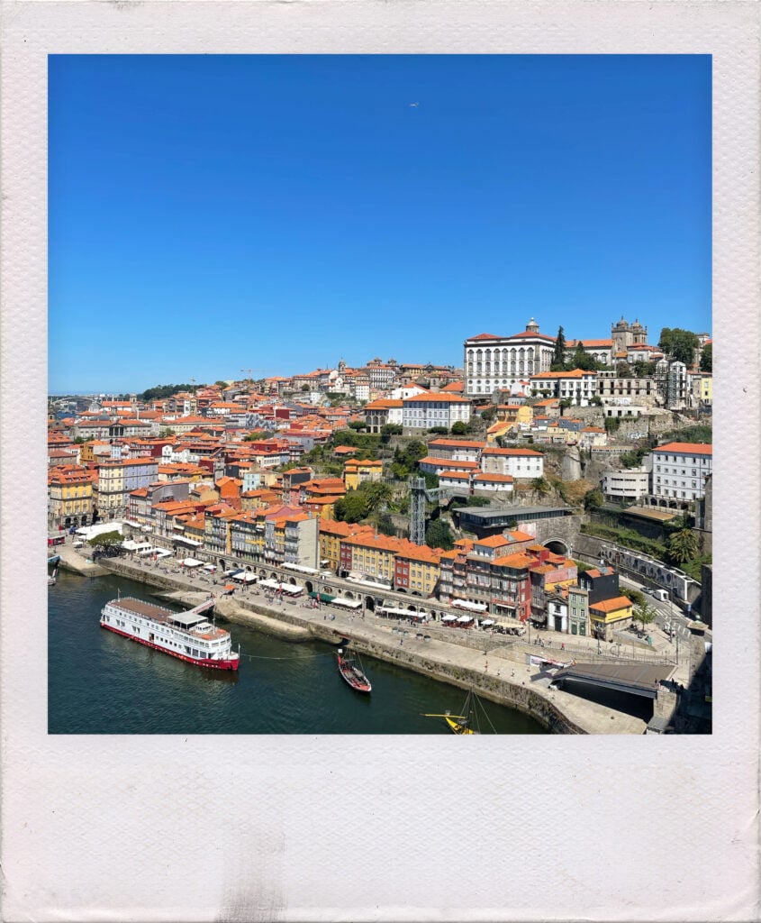 An aerial view of the Porto waterfront with colorful buildings, red-tiled roofs, and boats on the Douro River under a clear blue sky.