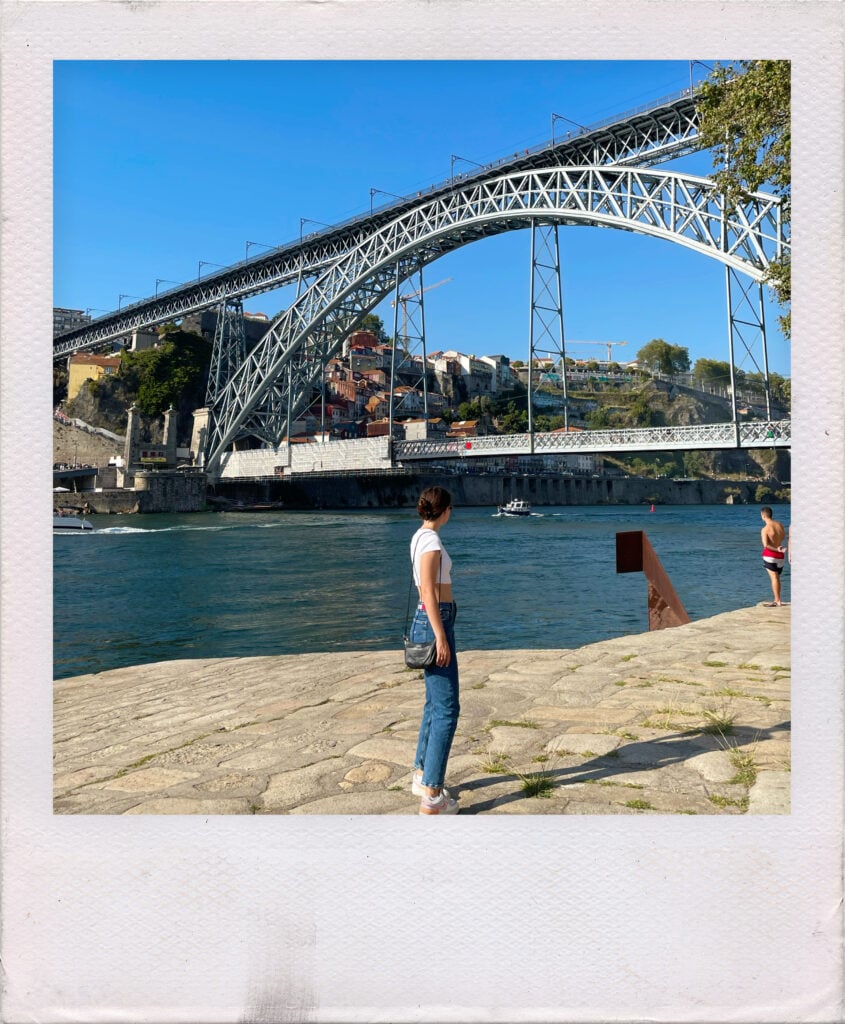 A woman stands on a stone embankment looking toward the Dom Luís I Bridge over the Douro River in Porto.