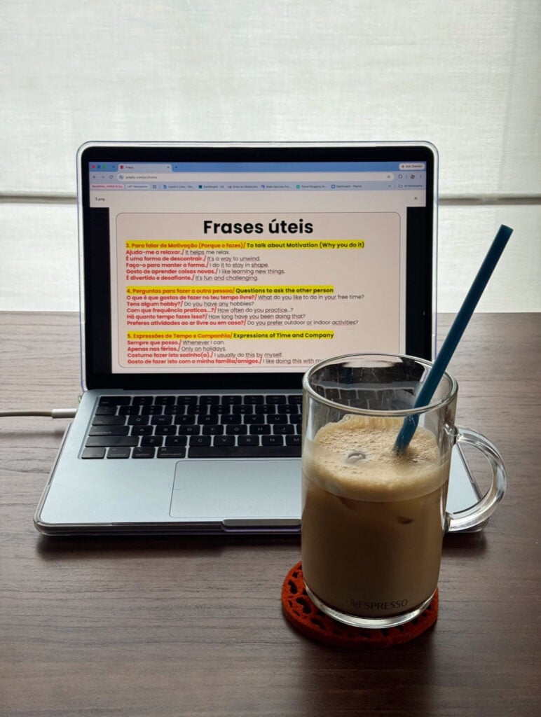A laptop screen displaying a Portuguese and English language learning document, positioned next to a glass of iced coffee with a blue straw.